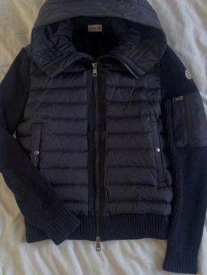 Mörkblå cardigan från Moncler - Snygg mörkblå pufferjacka från Moncler med quiltad front och rygg, stickade ärmar och muddar samt en stor huva. Jackan har dragkedja framtill, två fickor med tryckknappar och en extra ficka på ärmen med Moncler-logga. Perfekt för kalla vinterdagar.