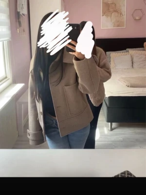 Beige ulljacka med stora fickor - Snygg beige jacka i ull med stora framfickor och bred krage. Jackan har en rak passform och långa ärmar med uppvikta muddar. Perfekt för dig som vill ha en stilren och trendig look.