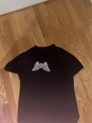 Svart t-shirt med vingar och text - Cool svart t-shirt med tryck av vita vingar och en liten rosa detalj på bröstet. På ryggen står det 'Angels' i stor vit text. Perfekt för dig som gillar streetwear och vill sticka ut lite extra.