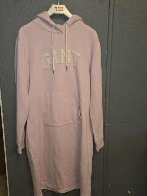 Ljusrosa hoodieklänning från GANT - Mysig ljusrosa hoodieklänning från GANT med broderad logga framtill. Har huva med dragsko, långa ärmar med muddar och en stor magficka. Perfekt för chill dagar hemma. Tillverkad i mjukt sweatshirtmaterial.