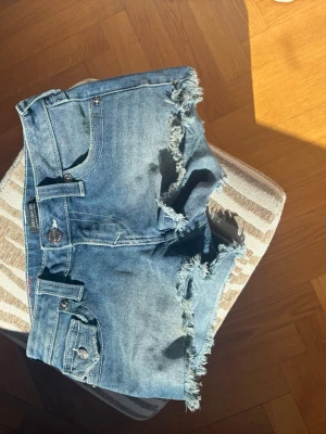 Blå jeansshorts från True Religion - Säljer ett par blå jeansshorts från True Religion med fransiga kanter och klassiska fickor både fram och bak. Shortsen har låg midja och är tillverkade i bomull. Perfekta för varma dagar och en avslappnad stil.