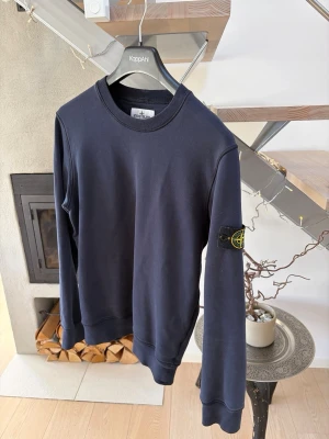 Mörkblå sweatshirt från Stone Island - Säljer en stilren mörkblå sweatshirt från Stone Island med klassisk rund hals och ikonisk logotyp-patch på vänster ärm. Tröjan har ribbade muddar vid ärmslut och nederkant. Köpt på NK för några år sedan.