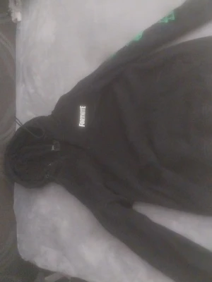 Svart Fortnite hoodie med tryck - Säljer en svart hoodie från Fortnite med vitt Fortnite-tryck på bröstet och coola gröna motiv på sidan. Hoodien har huva med dragsko och magficka framtill. Perfekt för dig som gillar gaming och vill ha en snygg och bekväm stil.