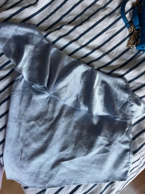 Ljusblå oneshoulder topp Gina Tricot - Snygg ljusblå oneshoulder topp från Gina Tricot med volangdetalj längs axeln. Toppens material har en lätt struktur och känns mjuk mot huden. Super fin nu till sommaren! 