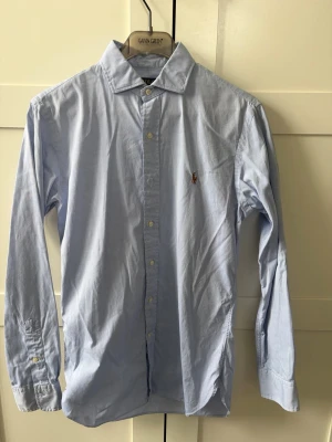 Ljusblå skjorta från Polo Ralph Lauren - Klassisk ljusblå skjorta från Polo Ralph Lauren i slim fit-modell. Skjortan har långa ärmar, knappar framtill och den ikoniska broderade loggan på bröstet. Tillverkad i mjuk bomull och passar perfekt till en stilren look.