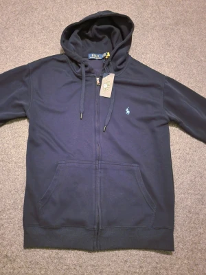 Mörkblå Polo Ralph Lauren zip up/ kofta - Snygg mörkblå hoodie från Polo Ralph Lauren med dragkedja och huva med snörning. Den är I storlek M kan även passa S. Tillverkad i 100% mjuk bomull. OBS: färgen ser lite dåligare ut på bilden men inte i verkligheten. Vid snabbaffär kan priset diskuteras och även frakta vid det första tillfället.