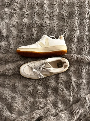 Veja V-12 vita sneakers med beige detaljer - Säljer ett par Veja V-12 sneakers i vitt läder och mesh med beige detaljer och gummisula. Klassisk låg modell med V-logga på sidan och snörning framtill. Perfekta för dig som gillar stilrena och trendiga sneakers.