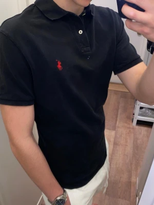 Polo Ralph Lauren (Svart piké) - Snygg svart piké från Polo Ralph Lauren med klassisk krage och knappar framtill. Ikonisk röd broderad logga på bröstet. Den är oanvänd och redo för en ny ägare! Pikén är storlek S men passar även M!