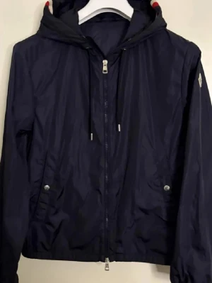 Mörkblå vindjacka från Moncler - Snygg mörkblå vindjacka från Moncler med huva och dragkedja framtill. Jackan har två fickor med tryckknappar, justerbar dragsko i huvan och Moncler-logga på ärmen. Lätt och smidig modell i glansigt material, perfekt för blåsiga dagar. Storlek 2 passar S/M. Självklart är den äkta!