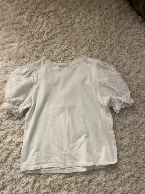 Vit t-shirt med puffärmar och broderi - Säljer en vit t-shirt från New Yorker med puffiga kortärmar i broderad bomull. Ärmarna har ett blommigt mönster och ger en romantisk vibe. Perfekt till jeans eller kjol för en söt och trendig look.