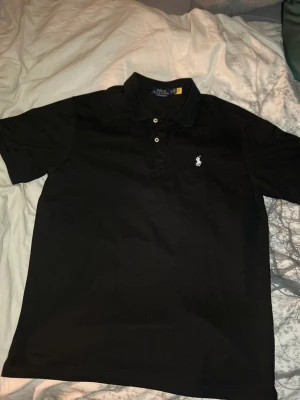 Svart pikétröja från Ralph Lauren - Klassisk svart pikétröja från Ralph Lauren med vit broderad logga på bröstet. Tröjan har krage, knäppning med två knappar och korta ärmar. Perfekt för en clean och stilren look.