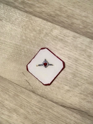Ring med röd hjärtformad sten - Snygg ring med en hjärtformad röd sten i mitten och små klara stenar längs bandet. Ringen har en elegant och romantisk design, perfekt för att sticka ut. Metallfärgen är silver. Kommer i en röd och vit ask.