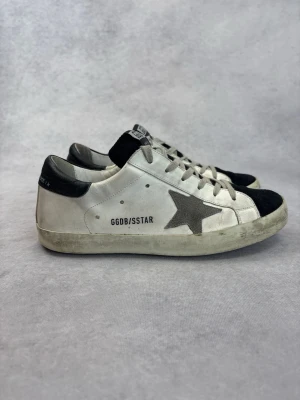 Golden Goose vita sneakers med stjärna - Snygga vita sneakers från Golden Goose med svart tå och häl, samt den ikoniska grå stjärnan på sidan. Skorna har vita snören och GGDB/SSTAR-tryck på sidan. Tillverkade i läder med detaljer i mocka för en cool kontrast.