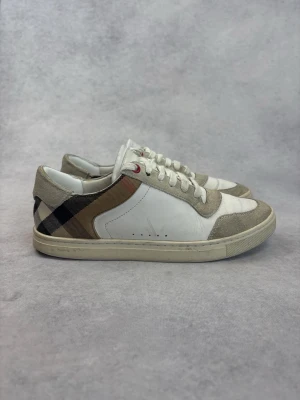 Burberry vita låga sneakers - Snygga vita sneakers från Burberry med klassiskt rutigt mönster i beige, svart och rött på sidan. Skorna har mockadetaljer i grått och vita skosnören. Tillverkade i en mix av läder och mocka för en exklusiv känsla. Perfekta för dig som vill ha en stilren och trendig look.
