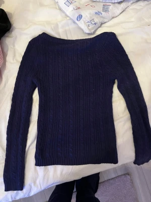 Mörkblå kabelstickad tröja - Mysig mörkblå kabelstickad tröja med lång ärm och rundad halsringning. Tröjan har ett klassiskt flätmönster och ribbstickade muddar vid halsen. Perfekt för kyliga dagar och enkel att matcha med jeans eller kjol.