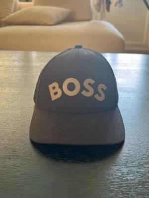 Svart keps från BOSS - Snygg svart keps från BOSS med stor vit logga framtill. Klassisk böjd skärm och justerbar baksida. Perfekt accessoar för en streetwear-look eller för att lyfta din outfit. Tillverkad i ett slitstarkt tygmaterial.