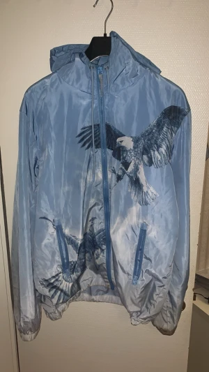 Trapstar Vindjacka - Snygg blå vindjacka med dragkedja och huva. Jackan har ett stort tryck av flygande örnar på framsidan och två fickor med dragkedja. Materialet är tunt och glansigt, perfekt för blåsiga dagar. Unik design som sticker ut.
