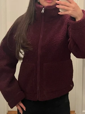 Vinröd teddyjacka med dragkedja - Helt ny med prislapp. Lager 157. Mysig vinröd teddyjacka med hög krage och hel dragkedja framtill. Jackan har två stora fickor och är gjord i fluffigt, mjukt material som håller dig varm. 