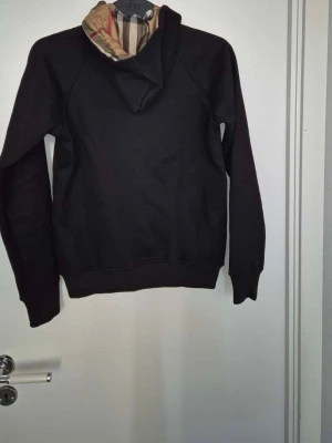 Svart hoodie från Burberry med dragkedja - Svart hoodie från Burberry med klassiskt rutigt foder i huvan och guldfärgad dragkedja framtill. Hoodien har snörning i huvan och en stor magficka. Tillverkad i mjuk bomull för en skön känsla.