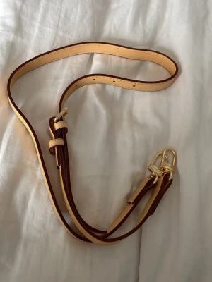 Justerbar axelrem i läder - Snygg axelrem i ljust och mörkbrunt läder med guldfärgade metalldetaljer. Remmen är justerbar och har klassiska sömmar längs kanterna. Perfekt för att byta ut eller uppgradera din väska med en stilren och tidlös look. Ny, i plast. 💫 Postar snabbt med en extra gåva vid snabbköp!