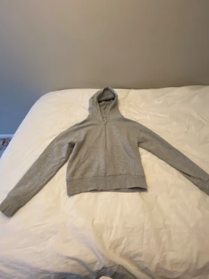 Grå zip hoodie med huva - En enkel och stilren grå hoodie med dragkedja framtill och två fickor. Hoodien har en klassisk huva och ribbade muddar vid ärmslut och nederkant. Perfekt för en avslappnad look och funkar året runt. Materialet känns mjukt och bekvämt, troligen bomullsblandning.