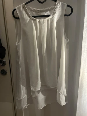 Vit ärmlös blus med pärldetaljer - Säljer en vit, flowy blus från Vero Moda. Blusen är ärmlös och har dubbla lager tyg för en luftig känsla. Rund halsringning med snygga pärldetaljer som ger extra stil. Perfekt till jeans eller kjol för en chic look.