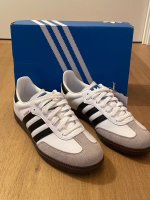 Adidas Samba vita och svarta sneakers - Klassiska Adidas Samba sneakers i vitt och svart med de ikoniska tre ränderna på sidan. Ovandelen är i vitt läder med grå mockadetaljer på tån och svart gummisula. Skorna har vit plös och vita skosnören samt blå Adidas-logga på plösen. Perfekt retrostil för streetwear.