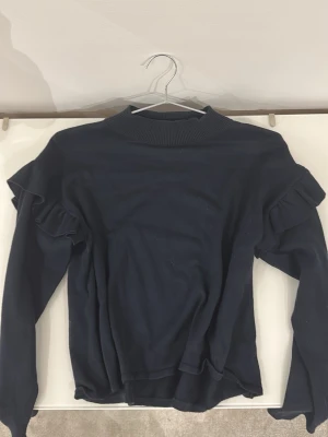 Mörkblå stickad tröja med volanger - Snygg mörkblå stickad tröja med volangdetaljer på axlarna och hög ribbad krage. Tröjan har långa ärmar och en mjuk, följsam passform som ger en trendig look. Perfekt för dig som gillar stilrena plagg med en twist.