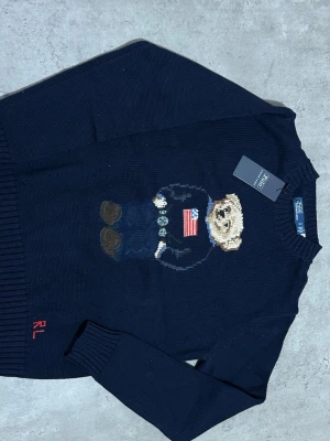 Mörkblå stickad tröja Polo Bear - Mörkblå stickad tröja från Polo Ralph Lauren med ikoniskt Polo Bear-motiv framtill. Tröjan har rund hals, ribbstickade muddar och RL-broderi vid nederkanten. Perfekt för dig som gillar klassisk och lekfull stil.