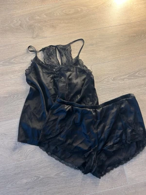 Svart satinpyjamas med spetsdetaljer - Snygg svart pyjamas i satin med linne och shorts. Linnet har tunna axelband och spets vid ringningen, medan shortsen har elastisk midja och spets längs kanterna. Perfekt för en lyxig och bekväm känsla på kvällen.