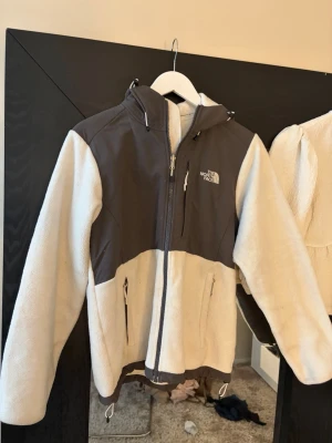 The North Face fleecejacka vit/grå - Säljer en snygg fleecejacka från The North Face i vit och mörkgrå färg. Jackan har dragkedja framtill, två fickor med dragkedja och huva. Perfekt för lager-på-lager och chill vardagsstil. Materialet är mjukt och värmande fleece.