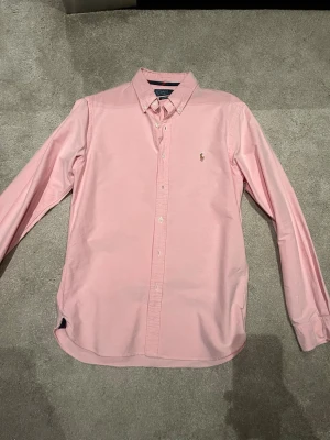 Rosa skjorta från Ralph Lauren - Klassisk rosa skjorta från Ralph Lauren med button-down krage och broderad logga på bröstet. Skjortan har lång ärm, normal passform och är tillverkad i mjuk bomull. Perfekt till jeans eller chinos för en clean och stilren look.