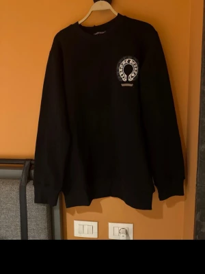 Svart sweatshirt från Chrome Hearts - Svart sweatshirt från Chrome Hearts med rund halsringning och ribbade muddar. Bröstet pryds av ett vitt tryck med Chrome Hearts-loggan. Perfekt för en avslappnad och cool stil.