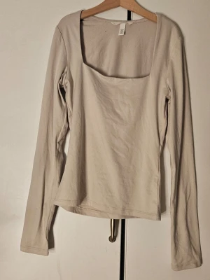 Beige långärmad topp - Stilren beige långärmad topp med fyrkantig ringning. Tillverkad i mjuk bomull som känns skön mot huden. Enkel och tidlös design utan mönster eller detaljer. Den är ny utan prislappar, men tvättad en gång för försäljning