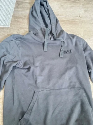 Grå hoodie från EA7 Emporio Armani - Snygg grå hoodie från EA7 Emporio Armani med klassisk huva och dragsnören. Stor magficka framtill och diskret logga på bröstet. Tillverkad i mjukt bomullsmaterial som känns skönt mot huden. Perfekt för en avslappnad och trendig look.