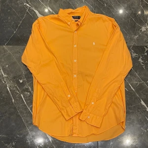 Orange linneskjorta från Polo Ralph Lauren - Klassisk orange skjorta från Polo Ralph Lauren med vit broderad logga på bröstet. Skjortan har lång ärm, knappar framtill och en tidlös krage. Perfekt för dig som vill sticka ut med färg och ändå hålla det stilrent. Använd enstaka gånger
