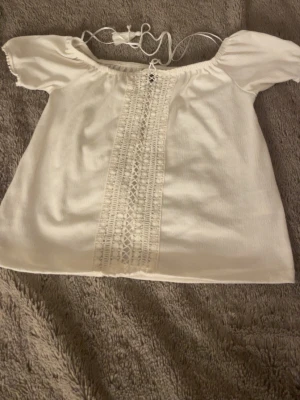 Vit offshoulder topp med spetsdetalj - Söt vit offshoulder topp med korta puffärmar och dekorativ spets längs mitten framtill. Toppen har snörning vid halsen och är gjord i ett lätt tyg som passar perfekt till sommaren.