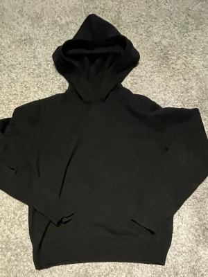 Svart hoodie med huva - En klassisk svart hoodie med huva och långa ärmar. Hoodien har en enkel och stilren design utan synliga loggor eller tryck. Perfekt för en avslappnad och cool look. Materialet ser ut att vara mjukt och bekvämt, troligen bomull eller bomullsblandning.