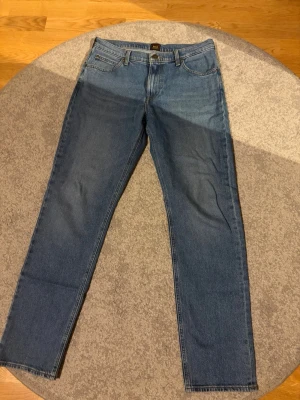 Blå raka jeans från Lee - Klassiska blå jeans från Lee med rak passform och fem fickor. Jeansen har normal midja, gylf med dragkedja och knapp samt Lee-logga på bakfickan. Tillverkade i denim med en snygg tvättad look.