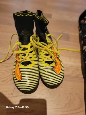  Fotbollsskor med högt skaft - Säljer ett par fotbollsskor med högt skaft i svart och gult, med gula snören och coola orangea detaljer på sidan. Skorna har ett randigt mönster.