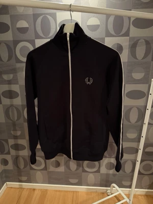 Svart zip hoodie från Fred Perry - Snygg svart hoodie från Fred Perry med vit dragkedja och vit rand längs ärmarna. Klassisk broderad lagerkrans-logga på bröstet. Tillverkad i mjukt och bekvämt material, perfekt för en avslappnad stil. Två fickor framtill och hög krage.