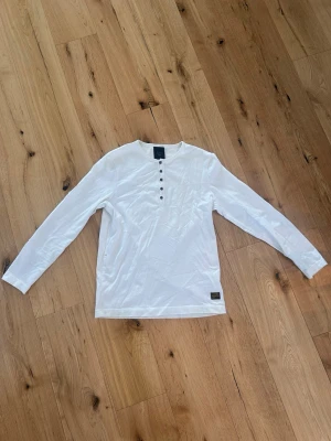 G-Star RAW vit långärmad Henley - Vit långärmad henley från G-Star RAW Correctline med bruna knappar framtill och diskret broderad logga på bröstet. Tillverkad i mjuk bomull för en avslappnad stil.