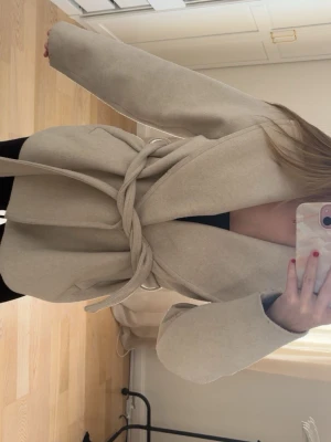 Beige kappa från Zara med knytskärp - Stilren beige kappa från Zara med bred krage och knytskärp i midjan. Kappan har en rak passform, två fickor framtill och är tillverkad i ett mjukt tyg.