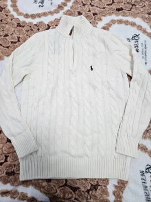 Vit kabelstickad tröja Polo Ralph Lauren - Kabelstickad vit tröja från Polo Ralph Lauren med hög krage och half zip-dragkedja. Tröjan har lång ärm, ribbade muddar och den klassiska broderade loggan på bröstet. Perfekt för lager på lager under kyliga dagar.