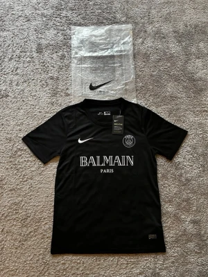 Nike x Balmain PSG t-shirt - Snygg svart t-shirt från Nike i samarbete med Balmain och PSG. T-shirten har korta ärmar, vit logga och text framtill samt är tillverkad i ett lätt och andningsbart polyestermaterial med Dri-FIT teknologi. Perfekt för dig som gillar streetwear och sportig stil.