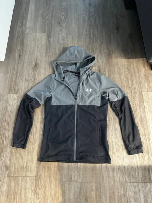 Grå och svart vindjacka Under Armour - Snygg tvåfärgad vindjacka från Under Armour i grått och svart. Jackan har huva, hel dragkedja framtill och två sidofickor. Skicket är som nytt. Passar S/M