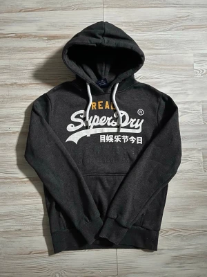 Mörkgrå Superdry hoodie med tryck - Snygg mörkgrå hoodie från Superdry med stor vit logga och japansk text på bröstet🙌🏻 inga defekter elle nåt som syns på hoodien. Kom gärna med bud och skriv gärna vid funderingar🤩