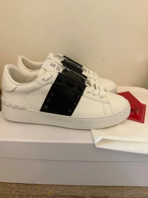Valentino Garavani vita sneakers med nitar - Snygga vita sneakers från Valentino Garavani med svart band och coola svarta nitar på sidorna. Skorna har en klassisk låg profil och är tillverkade i läder med vita snören och detaljerad logga på plösen. Perfekta för dig som vill sticka ut med stil.