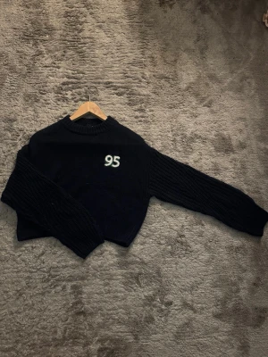 Mörkblå stickad tröja från Divided - Mysig mörkblå stickad tröja med broderad siffra '95' i vitt på bröstet. Tröjan har grovstickade ärmar, ribbad halsringning och muddar. Perfekt för dig som gillar en enkel men cool look. 