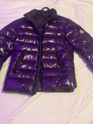 Lila glansig dunjacka från Moncler - Säljer en lila dunjacka från Moncler med glansigt yttermaterial och quiltad design. Jackan har hög krage, två dragkedjeförsedda fickor fram och Moncler-logga på ärmen. Perfekt för kalla vinterdagar och riktigt snygg att bära till det mesta. Dragkedja är defekt och ett jack i höger ärm     Mötas för köpas (Stockholm)